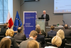 Wiceminister MSZ Jacek Czaputowicz przemawia z mównicy w auli KSAP. Widoczni siedzący uczestnicy konferencji. Obok mównicy baner KSAP i flagi polska i unijna.