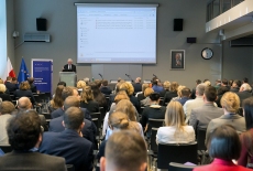 Wiceminister MSZ Jacek Czaputowicz przemawia z mównicy w auli KSAP. Widoczni siedzący uczestnicy konferencji. Obok mównicy baner KSAP i flagi polska i unijna. W tle duży ekran.