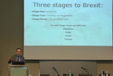 Prowadzący wykład stoi przy mównicy, obok na dużym ekranie wyświetlona prezentacja: Three stages to Brexit