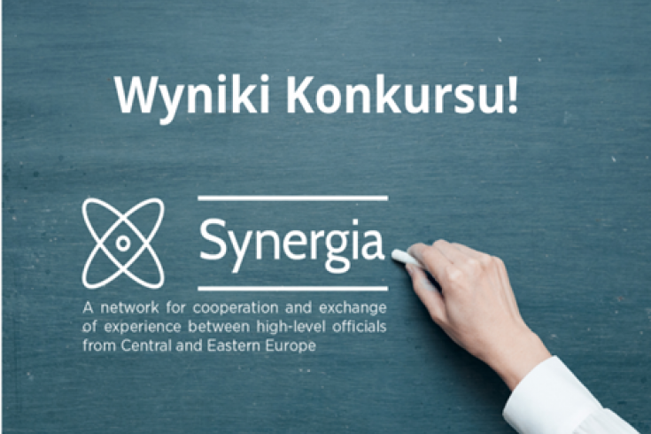 dłoń pisząca kredą na tablicy. napis: wyniki konkursu! Synergia